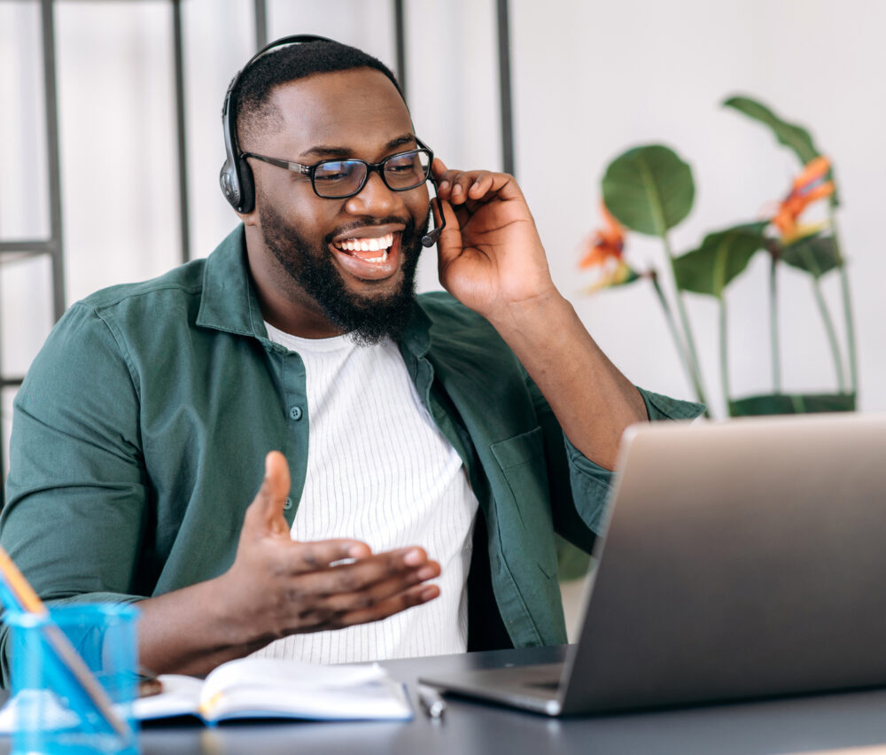 Friendly smiling African American guy, freelancer, student, confident call center operator in a headset, or ceo, uses laptop for video conference with coworkers or clients, telecommunication concept Friendly smiling African American guy, freelancer, student, confident call center operator in a headset, or ceo, uses laptop for video conference with coworkers or clients, telecommunication concept